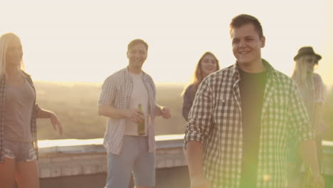 A-young-man-in-a-gray-plaid-shirt-and-black-t-shirt-moves-in-dance-with-his-friends.-He-enjoys-and-has-fun-with-beer-and-music-on-the-roof.